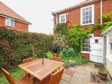 A garden patio with a wooden table and chairs surrounded by plants and a brick house with an open white door at Winkle Cottage Aldeburgh