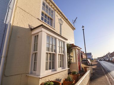 Exterior view of a yellow brick house with white framed windows and terracotta pots along the wall at Winkle Cottage Aldeburgh