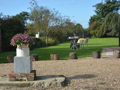 A garden with a flower pot on a stone pedestal tree stumps in gravel and a table with chairs on grass at Rookyards Spexhall Near Halesworth
