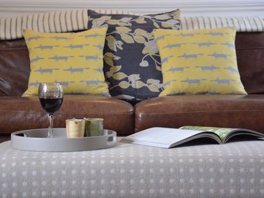 A brown leather sofa with two yellow patterned cushions and one dark floral cushion a gray tray with two small cups and a glass of red wine and an open book on a polka dot covered ottoman at Rookyards in Spexhall Near Halesworth