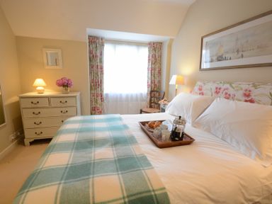 A bedroom with a bed covered with a check blanket and a tray with coffee and croissants on it a chest of drawers with a lamp and flowers near a window with floral curtains at 2 The Bays Thorpeness
