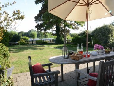 An outdoor patio with a wooden table and chairs under an umbrella overlooking a garden with a lake in the background at 2 The Bays Thorpeness