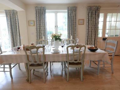 A dining room with a table set for a meal and chairs by windows with curtains at 2 The Bays Thorpeness