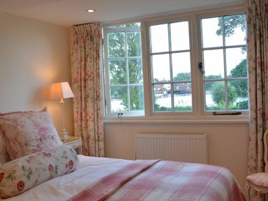 A bedroom with a bed covered in floral and plaid bedding next to a window with floral curtains and a lamp on a bedside table at 2 The Bays Thorpeness