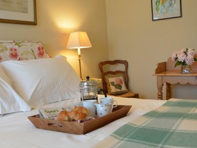 A bedroom with a bed with white sheets and a floral headboard a wooden chair with a floral cushion a wooden table with flowers and a tray with coffee and croissants on the bed at 2 The Bays Thorpeness