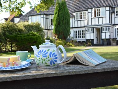A table outside with a teapot, a cup, a plate of snacks, and an open book in front of a large house at 2 The Bays Thorpeness
