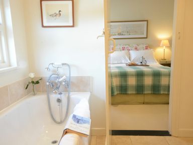 A bathroom with a bathtub and a book with a white rose next to it leading to a bedroom with a bed with a green plaid blanket and a table lamp at 2 The Bays Thorpeness