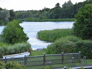 A lake surrounded by trees and plants with a small boat on the shore and chairs near a fence at 2 The Bays Thorpeness
