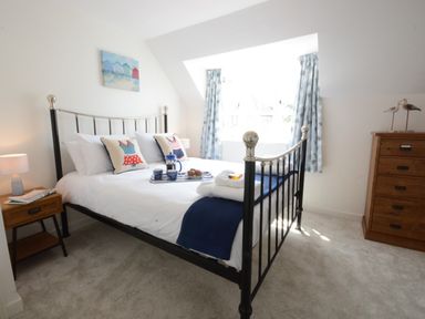 A bedroom with a metal bed frame bedside table with lamp wooden chest of drawers and window with curtains at 4 The Dunes Thorpeness