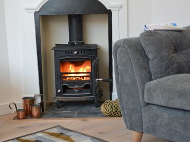 A wood burning stove with fire inside and copper containers next to a gray sofa at 4 The Dunes in Thorpeness