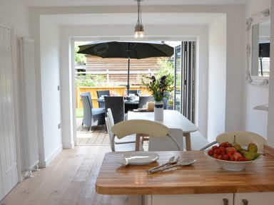 A kitchen view with a wooden countertop and bowl of fruit a dining table with chairs and flowers and an outdoor dining area with black umbrella and chairs at 4 The Dunes Thorpeness