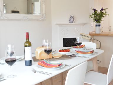 A dining table with wine glasses bread and tomatoes near a fireplace with flowers on a counter at 4 The Dunes Thorpeness