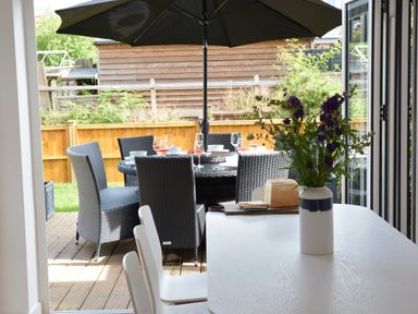 A garden dining set with a parasol on a decked patio and a table with a vase of flowers indoors at 4 The Dunes Thorpeness