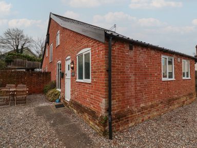 An exterior view of a brick building with a white door and windows and an outdoor wooden table set on gravel at 1 The Old Coach House Huntingfield Near Laxfield