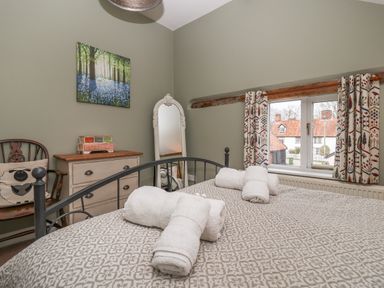 A bedroom with a bed and rolled towels on it a dresser a chair with a cushion a standing mirror and a window with patterned curtains at 1 The Old Coach House Huntingfield Near Laxfield