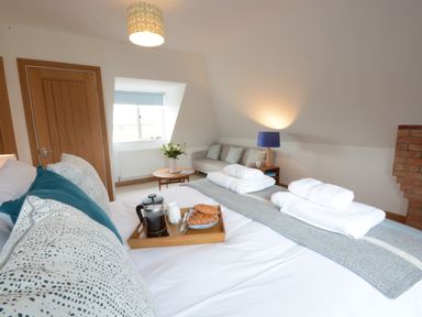 A bedroom with a bed set for breakfast, folded towels, a sofa, a side table with a lamp, a round coffee table with flowers, and a window at Ash Tree Cottage Aldringham