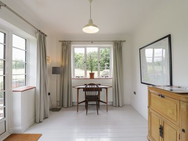 A room with white walls and floor with a wooden desk and chair near a window with curtains and a floor lamp beside the desk
