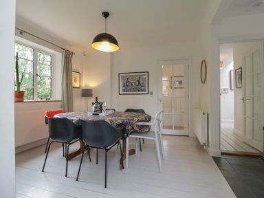 A dining room with a table covered by a patterned cloth surrounded by six chairs next to a window with curtains and a lamp on the wall at Ash Tree Cottage Aldringham