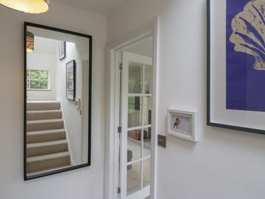 A hallway with a large mirror reflecting a staircase and framed pictures next to a glass-paneled door at Ash Tree Cottage Aldringham