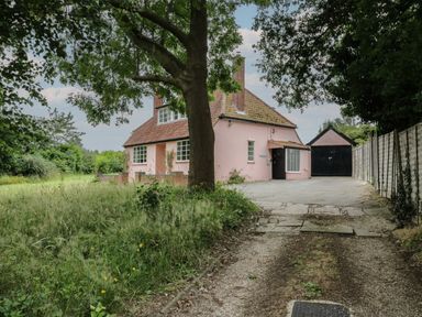A pink house with a chimney and a black garage surrounded by trees and a grassy area at Ash Tree Cottage Aldringham