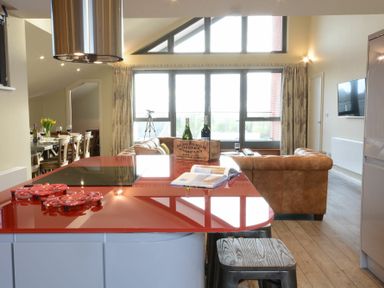 An open plan living area with a red kitchen island, brown sofas, large windows, and a dining table with chairs at King George Penthouse in Aldeburgh