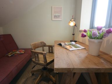 A wooden desk with a chair lamp an open book and flowers next to a window and a red sofa with books in a room at King George Penthouse in Aldeburgh
