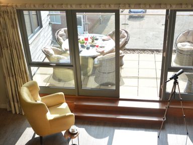 A room with a yellow armchair next to a small table and a telescope by glass doors leading to a patio with a round table and cushioned wicker chairs at King George Penthouse in Aldeburgh