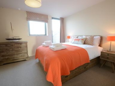 A bedroom with a double bed covered with an orange blanket and white towels bedside tables with lamps and a wooden dresser with a model sailboat at King George Penthouse in Aldeburgh