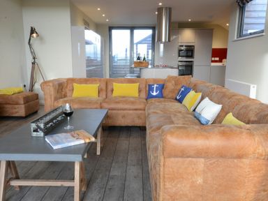 A living room with a large brown sectional sofa with yellow and blue cushions and a grey table with a glass of wine and a book at King George Penthouse in Aldeburgh