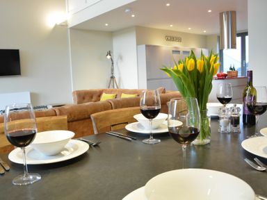 A dining table set with wine glasses, white bowls, and a vase of yellow flowers in a room with a brown sofa and a kitchen in the background at King George Penthouse in Aldeburgh