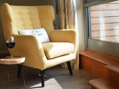 A yellow armchair with a white cushion and a glass of red wine on a small wooden table next to a window at King George Penthouse in Aldeburgh