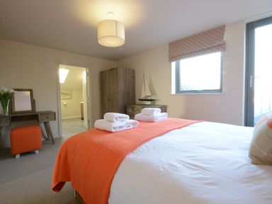 A bedroom with a bed covered in white and orange linens two towels on the bed a wooden desk with a mirror and a vase of flowers an orange stool a wooden wardrobe and dresser a model sailboat a window and a door at King George Penthouse in Aldeburgh