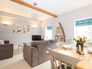A living room with gray sofas a coffee table a TV a boat-shaped bookshelf and a dining table with wine bottles and flowers at Saffron Blythview in Blythburgh near Southwold