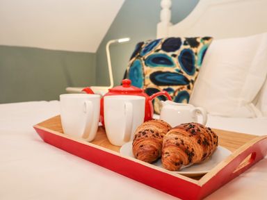 A tray with croissants mugs red teapot and milk jug on a bed with cushions in a bedroom at Saffron Blythview in Blythburgh near Southwold