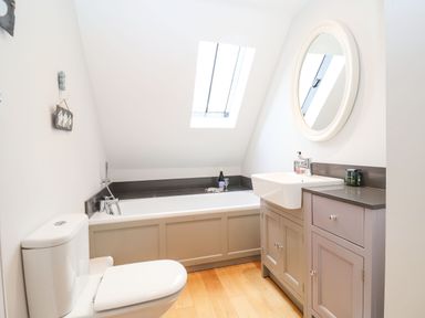 A bathroom with a toilet, bathtub under a skylight window, a sink with a cabinet, and a round mirror above the sink at Saffron, Blythview in Blythburgh near Southwold