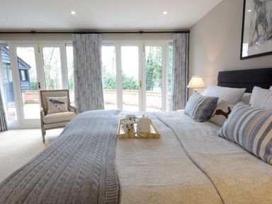 A bedroom with a large bed, striped pillows, a chair with a bird cushion, and glass doors leading outside at Bird Cottage, Burgh Near Grundisburgh