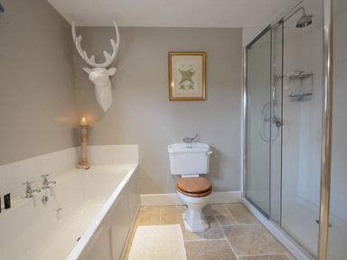 A bathroom with a bathtub, toilet with wooden seat, framed picture, glass shower enclosure, and decorative deer head on wall at Bird Cottage Burgh Near Grundisburgh