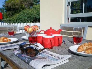An outdoor wooden table set with red mugs a red teapot glasses of red drink plates with croissants a bowl of apples and a newspaper with sunglasses at 1 South Cottages in Thorpeness