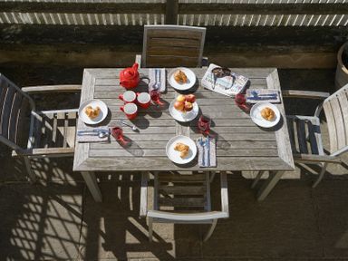 An outdoor wooden table set with four plates containing croissants four glasses with red drink a red teapot three red mugs a bowl of apples four sets of cutlery on napkins and a newspaper with sunglasses at 1 South Cottages in Thorpeness