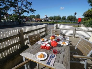An outdoor wooden table set with croissants apples red teapot cups and glasses on a patio with wooden chairs and white picket fence at 1 South Cottages in Thorpeness