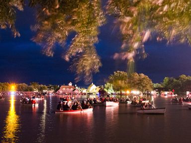 People in boats on a river at night with buildings and trees illuminated in the background at 1 South Cottages in Thorpeness