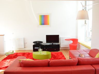 A living room with a red sofa a green coffee table a red rug and a TV on a black stand with a rainbow artwork on the wall at Parrotfish Bay in Southwold