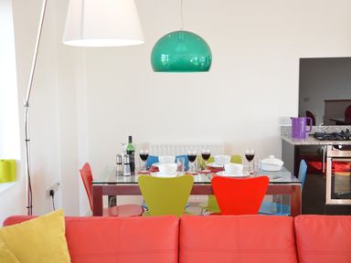 A dining area with a glass table set for four with colorful chairs and a green pendant light above at Parrotfish Bay Southwold