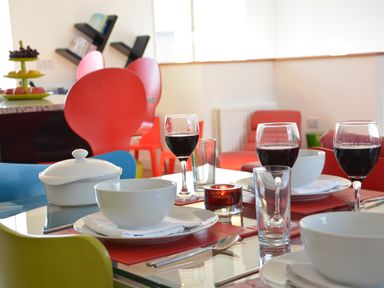 A dining table set with white bowls plates wine glasses and cutlery in a room with colorful chairs at Parrotfish Bay in Southwold