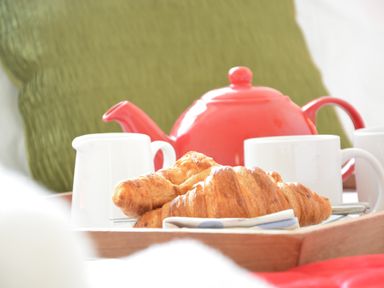 A tray with croissants cups a red teapot and a white jug on a bed with a green pillow at Parrotfish Bay Southwold