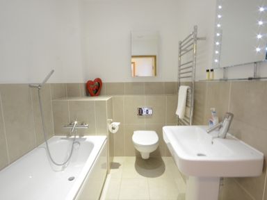 A bathroom with a bathtub handheld shower a wall mounted toilet sink mirror towel rail and heart shaped decoration at Parrotfish Bay Southwold