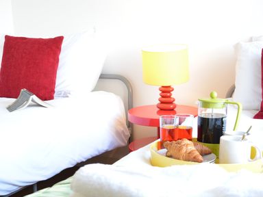 Two beds with white bedding and red pillows a bedside table with a red lamp and a breakfast tray with croissants drinks and a coffee pot at Parrotfish Bay in Southwold