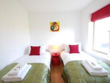 A bedroom with two single beds with green covers and red pillows a red bedside table with a lamp and a digital clock and a window with a red blind at Parrotfish Bay in Southwold