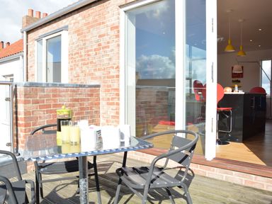 An outdoor patio with a metal table and chairs next to a sliding glass door leading to a kitchen with red chairs and yellow pendant lights at Parrotfish Bay in Southwold
