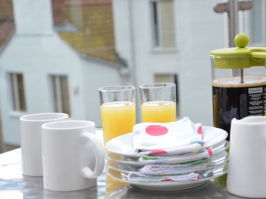Two white mugs two glasses of orange juice a stack of plates with colorful napkins and a french press coffee maker on a table